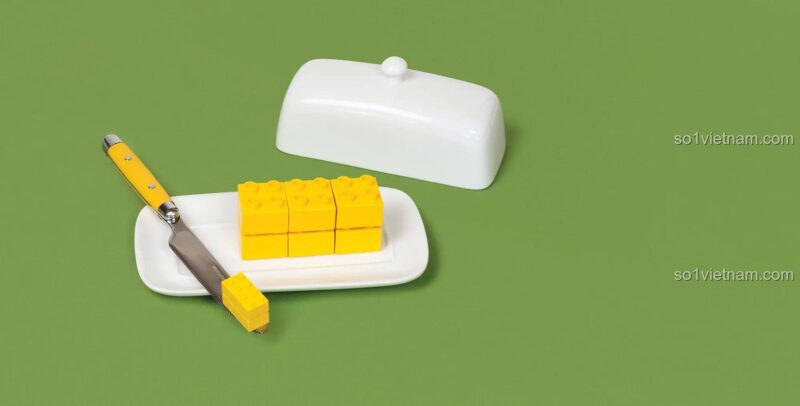 Sách ảnh LEGO Still Life with Bricks: The Art of Everyday Play của Chronicle, hình ảnh miếng bơ làm từ gạch LEGO® màu vàng trên đĩa, thể hiện nghệ thuật sắp đặt LEGO độc đáo.